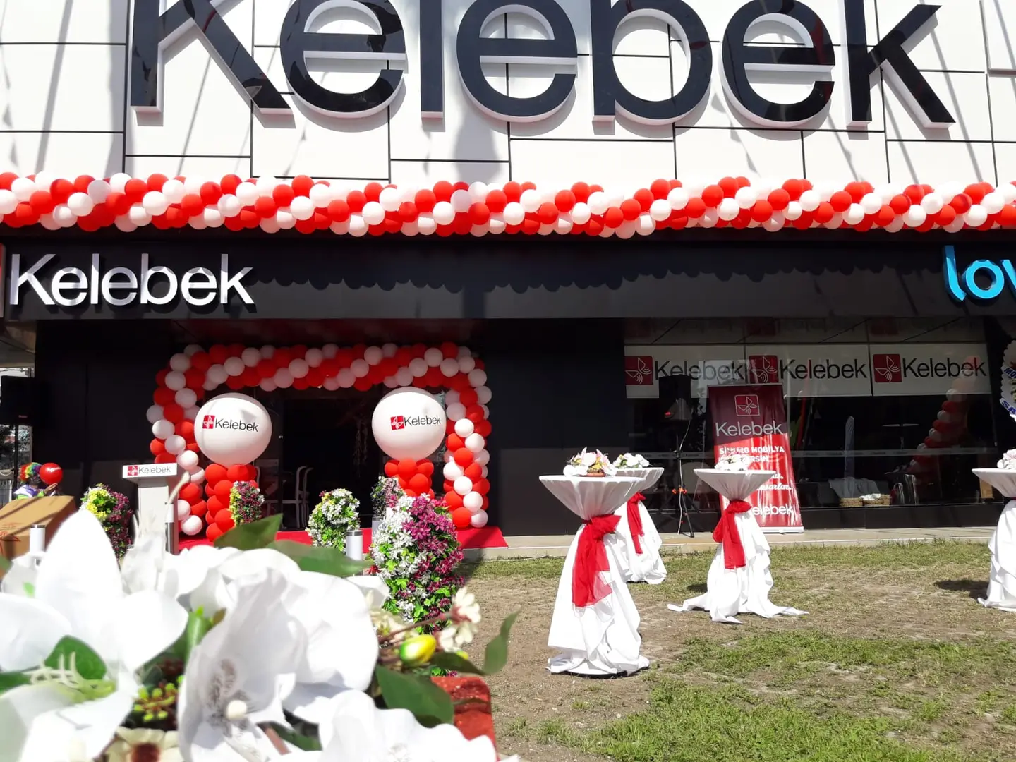 Mağaza cephesinde kırmızı-beyaz balon kemeri, çelenkler ve ikram masaları.